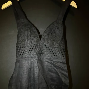 Svart klänning med spetsdetaljer -  svart långklänning med spetsdetaljer och snörning i ryggen. Klänningen har en v-ringad design och är ärmlös, vilket ger en stilren look. Perfekt för sommaren, storleken är XS men passar även i XXS