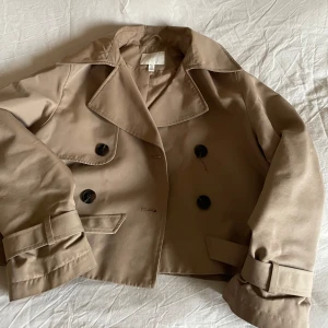 Beige trenchcoat - Snygg beige trenchcoat med dubbelknäppning och stora svarta knappar. Jackan har en klassisk krage och är croppad i modellen. Storlek xs men skulle säga mer S. Perfekt jacka till våren💕
