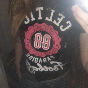 Svart t-shirt med tryck - Snygg svart t-shirt med stort tryck i vitt och rosa. Texten 'Celtic' och '88' ger en sportig känsla. Perfekt för en avslappnad stil.