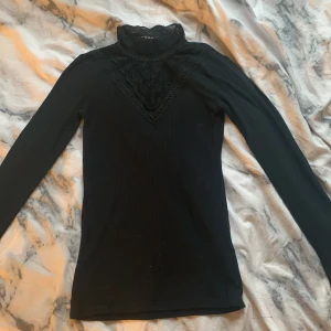 Svart långärmad topp från Name It - Säljer en elegant svart långärmad topp från Name It med spetsdetaljer vid halsen. Perfekt för att ge en stilren look. Toppen har en ribbad struktur som ger en skön passform.