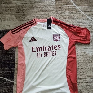  Fotbollströja från Lyon - Säljer en snygg vit och röd fotbollströja från Adidas med Olympique Lyon-logga och 'Emirates Fly Better' tryck. Tröjan har korta ärmar och är tillverkad i AEROREADY-material för optimal komfort. Perfekt för fotbollsfans och sportentusiaster!