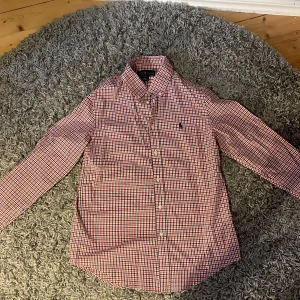 Rutig skjorta från Ralph Lauren - Snygg rutig skjorta från Ralph Lauren i rött, vitt och blått. Skjortan har långa ärmar och knappar framtill. Den klassiska loggan är broderad på bröstet. Perfekt för en stilren look! Nypris 2000