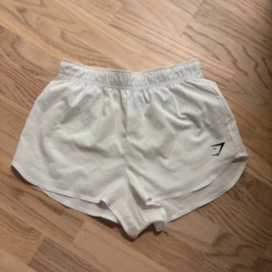 Vita träningsshorts från Gymshark - Snygga vita träningsshorts från Gymshark med elastisk midja och en liten logga på benet. Perfekta för gymmet eller löprundan. De har en bekväm passform och är gjorda i ett lätt material som andas.
