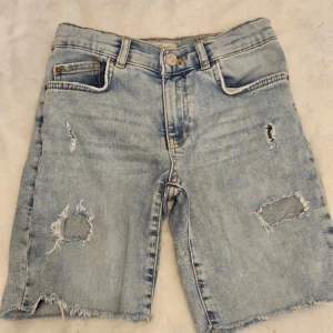 Ljusa jeansshorts från Zara - Säljer ett par ljusa jeansshorts från Zara med slitna detaljer och fransiga kanter. Shortsen har en klassisk femficksdesign och knappgylf. Perfekta för en avslappnad stil.