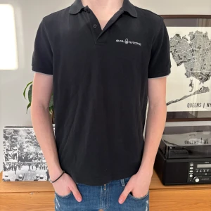 Svart pikétröja från Sail Racing - Säljer en stilren svart pikétröja från Sail Racing. Tröjan har en klassisk krage och korta ärmar, perfekt för en avslappnad look. Diskret logga på bröstet och en liten symbol på ryggen. Passar bra till jeans eller chinos.
