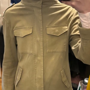 Grön  jacka med dragkedja - Säljer en stilren beige jacka med dragkedja och flera fickor framtill. Jackan har en normal passform och långa ärmar, perfekt för vår och höst. Enkla detaljer gör den lätt att matcha med olika outfits.