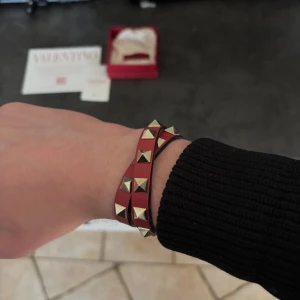 Rött armband med nitar från Valentino - Snyggt rött armband från Valentino med guldiga nitar. Armbandet har ett spänne i guld och är tillverkat i skinn. Perfekt för att ge en unik touch till din outfit.