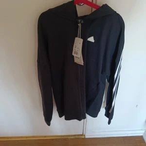 Svart huvtröja med dragkedja från Adidas - Säljer en svart huvtröja från Adidas med dragkedja och vita ränder längs ärmarna. Perfekt för en sportig look. Tröjan har en luva och är långärmad.