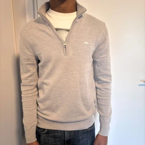 Grå tröja med dragkedja från J.Lindeberg - Snygg grå tröja från J.Lindeberg med halv dragkedja och ribbade muddar. Perfekt för en stilren look. Tröjan har en diskret logga på bröstet och är tillverkad i ett mjukt material.