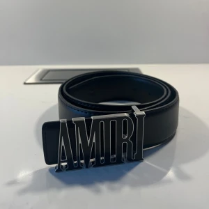 Svart bälte med AMIRI-spänne - Snyggt svart bälte från AMIRI med ett iögonfallande silverfärgat spänne som stavar ut märkesnamnet. Perfekt för att ge en stilren touch till din outfit.