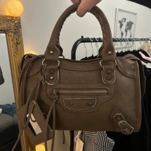 Balenciaga liknande väska - Snygg brun handväska från Balenciaga i skinn med dragkedja och dekorativa detaljer. Väskan har en praktisk storlek och kommer med en matchande tag. Bilderna beskriver skicket. Rostig runt ”nitarna” och metallen men annars är väskan superfin. Man kan även se de på bilderna tydligt 😊 Band ingår ej!