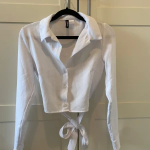 Vit croppad blus med knyt - Säljer en stilren vit croppad blus med knyt i midjan. Blusen har långa ärmar och knappar framtill. Perfekt för en trendig look!