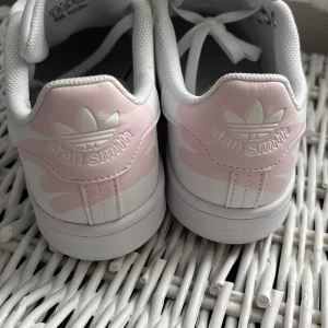 Vita och rosa Adidas Stan Smith sneakers - Säljer ett par klassiska vita Adidas Stan Smith sneakers med rosa detaljer. Skorna är i nästan nyanvänt skick, använt de två gånger ute (lite smutsig undersida) men annars har de bara legat i garderoben och samlat damm 