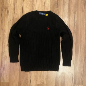 Svart stickad tröja från Ralph Lauren - Säljer en stilren svart stickad tröja från Ralph Lauren med det klassiska röda logotypbroderiet på bröstet. Tröjan har en ribbad struktur och långa ärmar, perfekt för kyligare dagar. Passar både till vardags och mer uppklädda tillfällen.