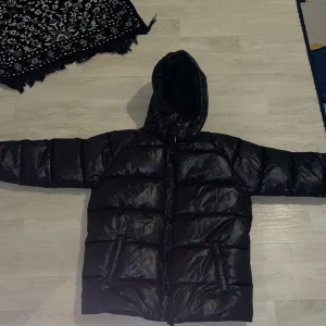 Svart pufferjacka med huva - Säljer en svart pufferjacka med huva. Jackan har en glansig yta och är perfekt för kyliga dagar. Den har en dragkedja framtill och långa ärmar. Perfekt för att hålla värmen under vintern. Storleken är 170 cm passar bra på folk lite längre eller lite kortare