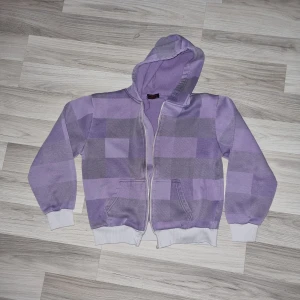 Lila  rutig hoodie - Säljer en lila rutig hoodie med dragkedja som tyvärr inte fungerar och behövs byta och fickor framtill. Den har en mysig huva och vita muddar vid ärmslut och nederkant. Perfekt för en avslappnad stil!