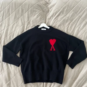 Svart tröja från Ami Alexandre Mattiussi - Säljer en stilren svart tröja från Ami Alexandre Mattiussi med ett rött hjärta och bokstaven A på bröstet. Tröjan har långa ärmar och ribbade muddar vid ärmslut och nederkant. Perfekt för en trendig look!