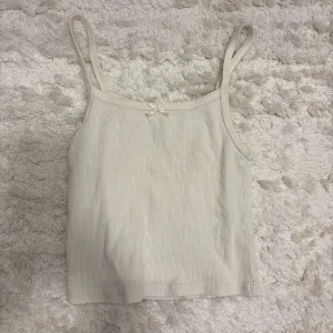 Vit ribbad linnetopp - Säljer en söt vit ribbad linnetopp med smala axelband och en liten rosett framtill. Perfekt för varma dagar eller som ett lager under en tröja.