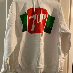Vit hoodie med 7UP-tryck från Vailent - Säljer en cool vit hoodie från Vailent med ett stort 7UP-tryck i rött och grönt på framsidan. Hoodien har en klassisk passform med långa ärmar och en praktisk ficka framtill. Perfekt för en avslappnad stil!