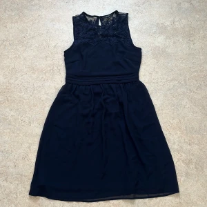 Mörkblå ärmlös klänning med spetsdetaljer - Snygg svart klänning från Veromoda med spetsdetaljer upptill och ärmlös design. Perfekt för en elegant look. Klänningen har en rund hals och knäppning i nacken. Materialet är lätt och luftigt med två lager tyg vilket ger en fin passform. Passande för skolavslutningen, dop, bröllop eller bara en helt vanlig sommardag. 