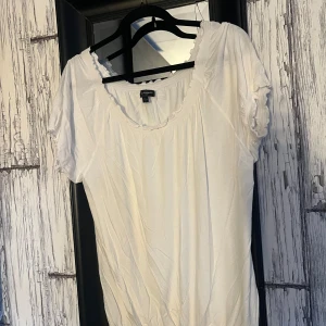 Vit blus från KappAhl - Säljer en söt vit blus från KappAhl med korta ärmar och fina volangdetaljer vid halsringningen. Perfekt för en avslappnad och stilren look. Blusen har en liten rosett framtill som ger en extra touch.