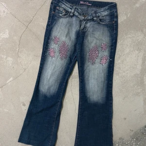 Blå jeans med broderade detaljer från Miss VS - Snygga blå jeans från Miss VS med unika rosa broderade detaljer på fram- och baksidan. Jeansen har en klassisk femficksdesign och en lätt utsvängd bootcut-stil. Perfekta för dig som vill ha något annorlunda i garderoben! Passar någon som är ungefär 165 cm! 