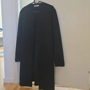 Svart kappa från Mango - Säljer en stilren svart kappa från Mango. Den har en enkel design utan krage och långa ärmar. Perfekt för en elegant look. Passar bra till både vardag och mer formella tillfällen.