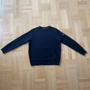 Svart stickad tröja från Canada Goose - Säljer en stilren svart stickad tröja från Canada Goose. Tröjan har långa ärmar och en rund halsringning. Perfekt för kyligare dagar med sin klassiska design och varma material.
