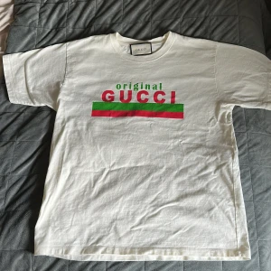 Vit t-shirt från Gucci - Säljer en vit t-shirt från Gucci med tryck i rött och grönt. T-shirten har korta ärmar och en klassisk rund halsringning. Perfekt för en stilren och lyxig look. Storleken är M