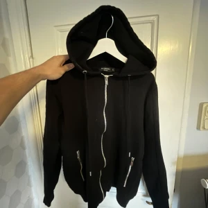 Svart hoodie med dragkedja - Säljer en svart hoodie med dragkedja och huva. Den har långa ärmar och två dragkedjefickor framtill. Perfekt för en casual look.