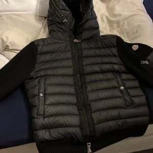 Svart dunjacka med huva - Snygg svart dunjacka från Moncler med huva och dragkedja. Jackan har långa ärmar och ribbade muddar. Praktiska fickor med knappar och en extra ficka på ärmen. Perfekt för kyliga dagar.