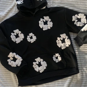 Svart hoodie med vita blommor från Denim Tears - Säljer en cool svart hoodie från Denim Tears med vita blommönster över hela tröjan. Hoodien har en klassisk passform med en stor ficka framtill och en bekväm huva. Perfekt för att sticka ut i mängden! Knappt använd