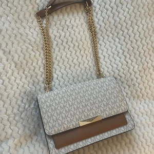 Axelväska från Michael Kors - Snygg axelväska från Michael Kors med ett stilrent mönster i beige och brunt. Väskan har en kedjerem i guld och en praktisk ficka på baksidan. 