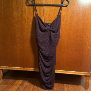 Lila kortklänning  - Säljer en elegant mörklila kortklänning från Windsor med glittrande detaljer. Klänningen har smala axelband och en dekorativ rosett framtill. Perfekt för en kväll ute eller en speciell tillställning. Exklusiv då jag köpte den i butik i Arizona + går ej att hitta på hemsidan och svår att få tag på någon annanstans, så passa på! 