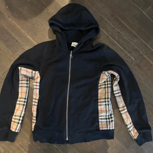Mörkblå hoodie från Burberry - Säljer en snygg mörkblå hoodie från Burberry med klassiskt rutigt mönster på insidan av ärmarna. Tröjan har dragkedja framtill och en bekväm huva. Perfekt för en stilren look!