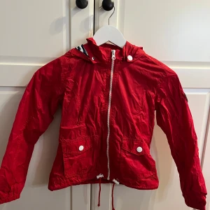 Röd vindjacka från Havana & Friends - Säljer en snygg röd vindjacka från Havana & Friends med randigt foder i huvan. Jackan har dragkedja och knappar framtill samt en cool patch på ärmen. Perfekt för blåsiga dagar!