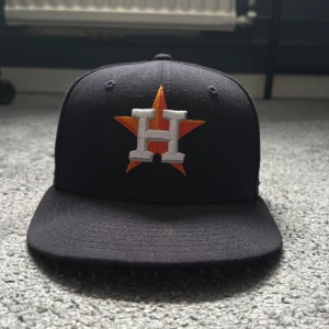 Marinblå keps med broderad logotyp - Snygg svart keps från New Era med en broderad logotyp i orange och vitt framtill. Kepsen har en platt skärm och MLB-logotypen baktill. Tillverkad i 100% polyester och passar perfekt för sportiga tillfällen.