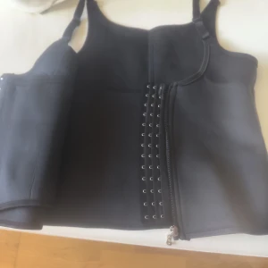 Svart korsett med dragkedja - Snygg svart korsett med dragkedja framtill. Perfekt för att ge en stilren look. Justerbara axelband för bästa passform.