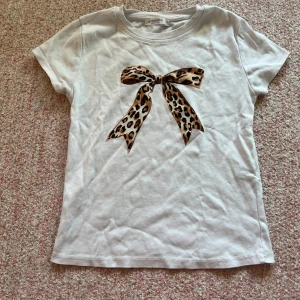 Vit t-shirt med leopardmönstrad rosett från Stradivarius - Säljer en söt vit t-shirt från Stradivarius med en stor leopardmönstrad rosett på framsidan. Perfekt för att ge din outfit en trendig touch. T-shirten har korta ärmar och en rund halsringning.