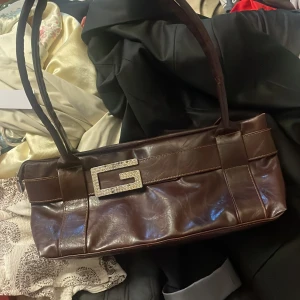 Brun axelväska från Guess i skinn - Säljer en brun avlång axelväska från Guess i glansigt skinn med dubbla axelband. Väskan har en stor, glittrig G-detalj framtill med strass och dragkedja upptill. Perfekt för dig som gillar statement-accessoarer och klassisk 00-talsstil.