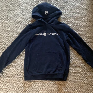 Mörkblå Sail Racing hoodie (160 cm) - Snygg mörkblå hoodie från Sail Racing med vit logga på bröstet och tryck på huvan. Klassisk känguruficka framtill och mjukt material som känns skönt mot huden. Perfekt för dig som gillar sportig och avslappnad stil.