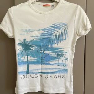 Vit t-shirt med tryck från Guess Jeans - Vit t-shirt från Guess Jeans med blått palmmotiv och stort frågetecken framtill. Trycket har en tropisk vibe och texten 'GUESS JEANS' . Klassisk passform och rund halsringning. Tillverkad i 100% bomull för en skön känsla.