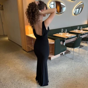 Svart långklänning från Desgin by Si - Supersnygg svart långklänning från Design by Si med djup v-ringning och draperad urringning fram. Klänningen är ärmlös och har en öppen rygg, figurnära passform och faller snyggt längs kroppen. Perfekt för bal eller festkvällar.