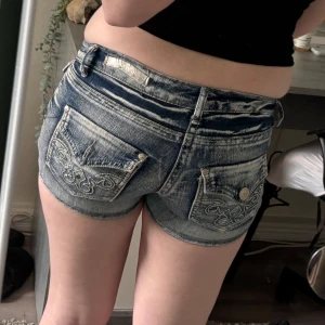 Lågmidjade jeansshorts med broderi - Snygga lågmidjade jeansshorts i blått med slitna detaljer och ljusa partier. Shortsen har broderade bakfickor med dekorativa sömmar och klassiska jeansknappar. Perfekta för sommaren och ger en cool, avslappnad vibe. Saknar några kristaller, säljer då de är för små.