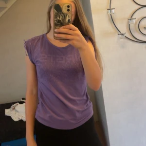 Lila sporttopp från Energetics - Lila sporttopp från Energetics med tryckt NRGTS-logga framtill. Toppen har korta ärmar med snygg rynk- och knyt-detalj på axlarna. Går att skjutstera längden på ärmarna. Materialet är mjukt och stretchigt, perfekt för träning eller aktiva dagar. Mönstret är diskret randigt i samma färgton.💜