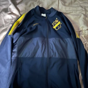 AIK träningströja - Snygg mörkblå träningsjacka från AIK med gula detaljer på ärmarna och klubbens emblem på bröstet. Jackan har dragkedja framtill och är tillverkad i ett sportig material. Royal edition priset är ej hugget i sten. Tröjan går ej att få tag i längre 