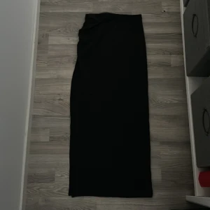 Svart långkjol med rynkdetalj - Svart långkjol i mjukt och stretchigt material med snygg rynkdetalj framtill i midjan. Kjolen har en enkel och clean look, perfekt om du gillar minimalistisk stil. Resår i midjan för extra komfort och smidig passform. Slits