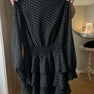 Svart prickig volangklänning - Supersöt svart kortklänning med vita prickar och volanger. Klänningen har långärmade ärmar med smock vid handlederna och markerad midja. Perfekt för dig som gillar en lekfull och trendig stil.