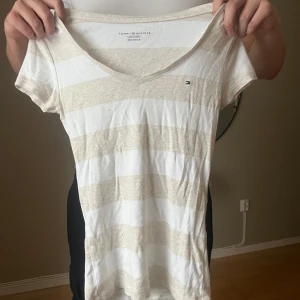 Randig v-ringad topp Tommy Hilfiger - Snygg v-ringad topp från Tommy Hilfiger i vitt och beige med breda ränder. Kortärmad modell med liten logga på bröstet. Perfekt för en avslappnad och stilren look.