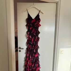 Lång röd och svart volangklänning - Supersnygg långklänning med tunna axelband och volanger över hela klänningen. Klänningen har ett rött och svart mönster och är gjord i ett lätt, transparent material som ger en cool och dramatisk look. Perfekt för dig som vill sticka ut med en unik stil.